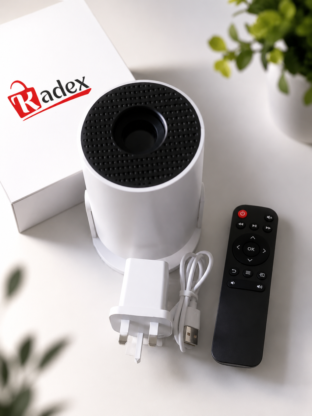 A white cylindrical Radex device with a black textured top, shown with its white charging adapter, USB cable, black remote control, and branded white box on a light tabletop.