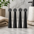 Four black electric toothbrush brush heads standing upright on a marble bathroom counter, with a soft towel, plant, and soap dispenser in a clean, modern background.