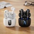 A side-by-side product shot of premium white and black wireless earbuds resting in their respective open charging cases, which show a digital battery life display, set against a softly blurred, warm oak desk background.