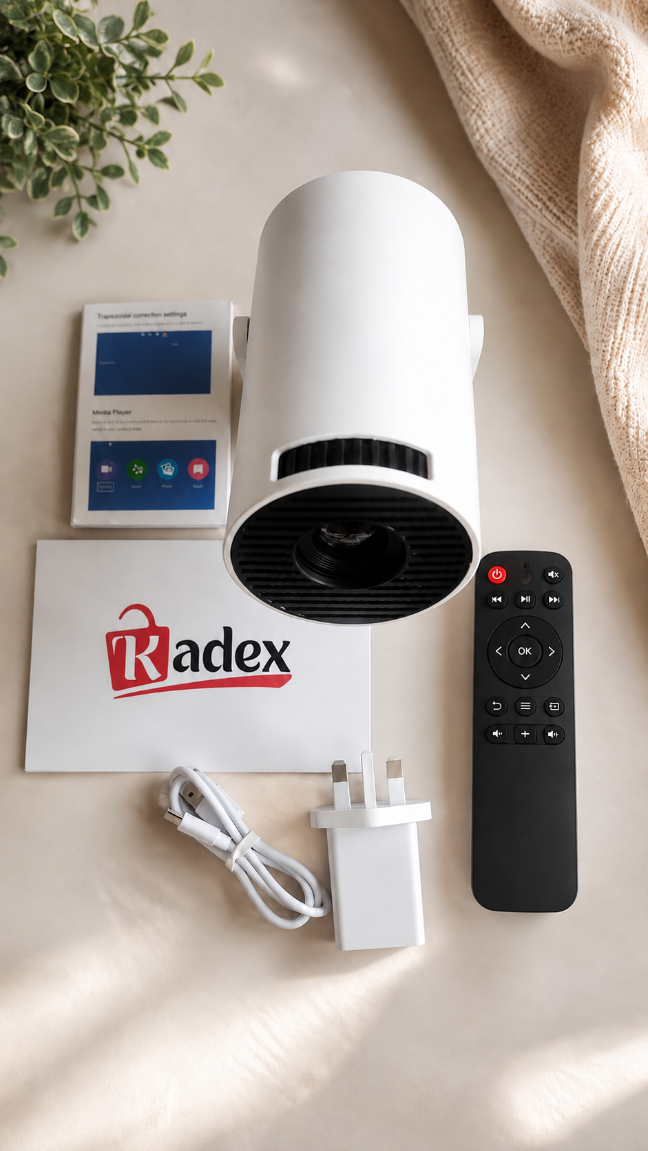 A white cylindrical Radex device with a black front grille, shown with a black remote control, white power adapter, charging cable, instruction booklet, and a small display screen on a beige surface with a plant and draped fabric.