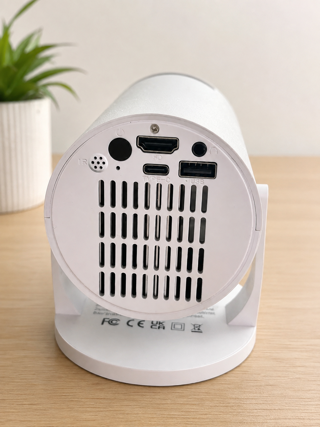 White cylindrical electronic device with ventilation grilles and multiple ports on the back, shown on a light wood tabletop with a blurred potted plant in the background.