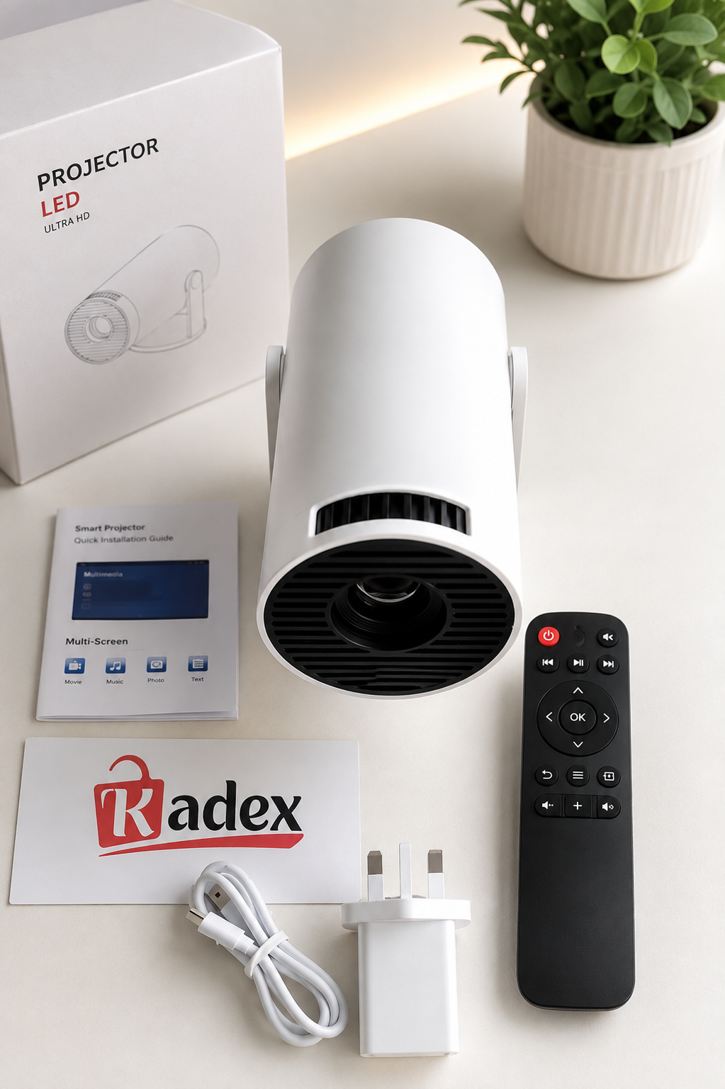 A white LED projector lamp with a black front lens opening, shown with its retail box, remote control, power adapter, USB cable, instruction booklet, and a Radex card on a light beige surface.