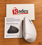 A white vertical optical mouse shown next to its user manual and a Radex brand card on a wooden surface.